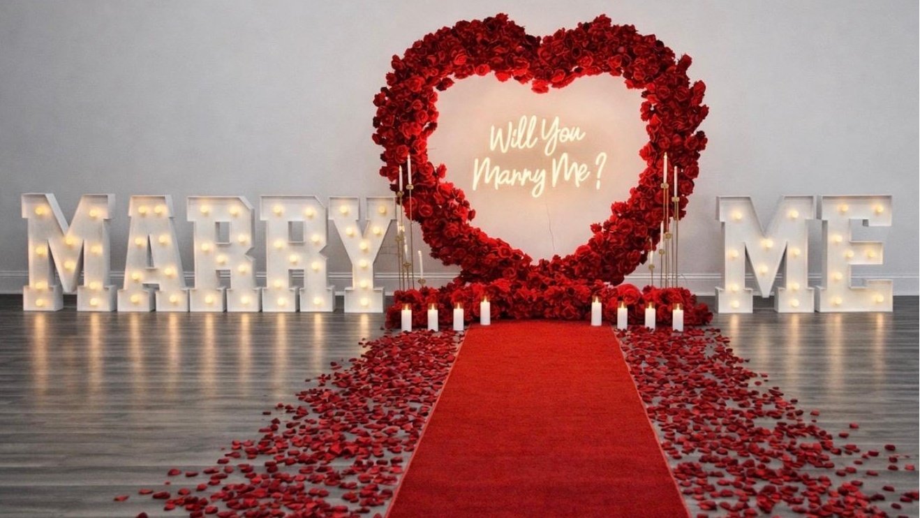 Red heart floral arch with Will You Marry Me neon sign, candles and rose petals in Chicago